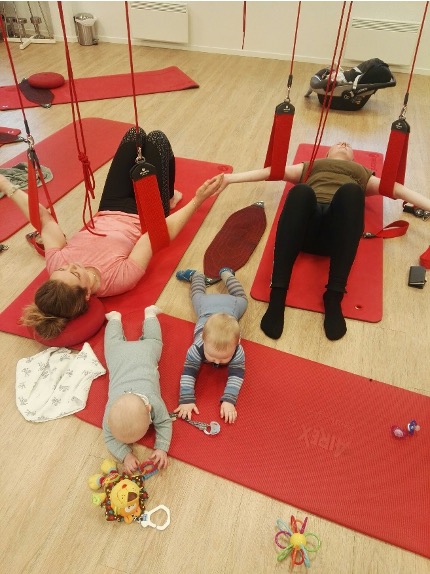 Two women exercising on mats with their legs in red Redcord slings while their babies play beside them